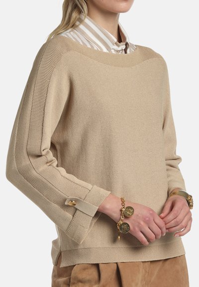 Pull beige en maille texturée, col large et poignets retroussés. Présente des accents à double bouton sur les manches, porté sur une chemise à rayures.