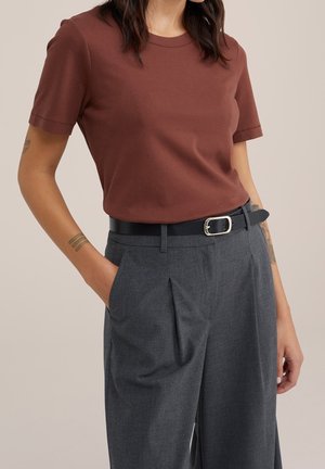 Kobieta ubrana w brązową koszulkę z krótkim rękawem, wpuszczoną w szare, plisowane spodnie z wysokim stanem i czarnym paskiem, stojąca z jedną ręką w kieszeni.