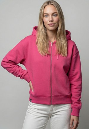 Junge Frau mit langen blonden Haaren trägt einen rosa Kapuzenpullover mit Reißverschluss und weiße Hose, steht mit einer Hand in der Hüfte vor einem grauen Hintergrund.