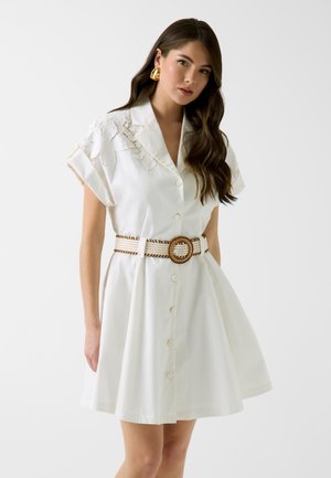 Młoda kobieta ubrana w białą sukienkę zapinaną na guziki z koronkowymi detalami na ramionach, pleciony pasek z okrągłą klamrą oraz złote kolczyki, stojąca na białym tle.