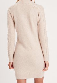 Robe en maille tricotée de couleur beige clair avec un col montant et des manches longues ; présente des détails en torsades le long des manches et une coupe ajustée.