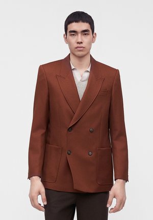 Ung mand iført en dobbeltradet rustbrun blazer over en lys skjorte, står mod en ensfarvet lys baggrund.