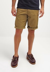 Shorts en coton beige avec des revers, associés à des chaussures de bateau en cuir marron foncé ornées de coutures blanches et d'une semelle texturée.