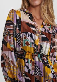 Robe à motifs géométriques multicolores en violet, jaune et marron. Elle présente une taille élastique, des manches longues et un décolleté en V. Tissu léger.