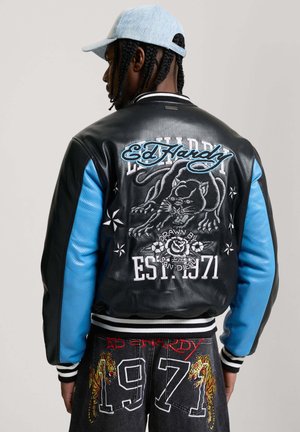 Persona que lleva una chaqueta Ed Hardy negra y azul y pantalones negros con tigres bordados y el texto "1971" en la parte trasera.