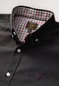 Camisa negra con botones, con cuello interior a cuadros, botones y una pequeña corona bordada en color burdeos en el pecho. Etiqueta visible en el interior.