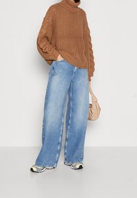Maglione a collo alto in maglia marrone con maniche testurizzate, abbinato a jeans in denim azzurro chiaro a gamba larga. Accessoriato con una borsa beige e sneakers.