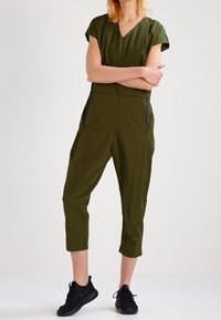 Mono verde olivo con mangas cortas, escote en V, pantalones cortos ajustados, bolsillos laterales y detalles en negro, hecho de un material ligero.