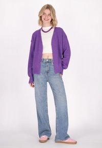Cárdigan de punto morado oversized con una textura suave, diseño de frente abierto, combinado con jeans de denim azul holgados y zapatillas rosas.