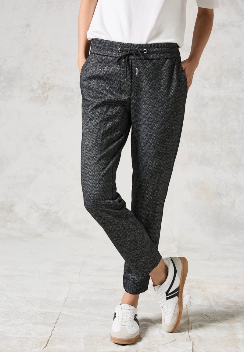 Personne portant un pantalon de jogging gris foncé motif chevrons avec taille à cordon et des baskets blanches à rayures noires, debout contre un mur clair texturé.