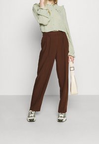 Green ruffled blouse, brown pleated trousers, and white handbag. Sneakers feature green and brown accents. Model stands against a neutral background.