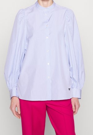 Kobieta ubrana w jasnoniebieską i białą koszulę w paski z guzikami, z bufkami na rękawach oraz w jasnoróżowe spodnie.