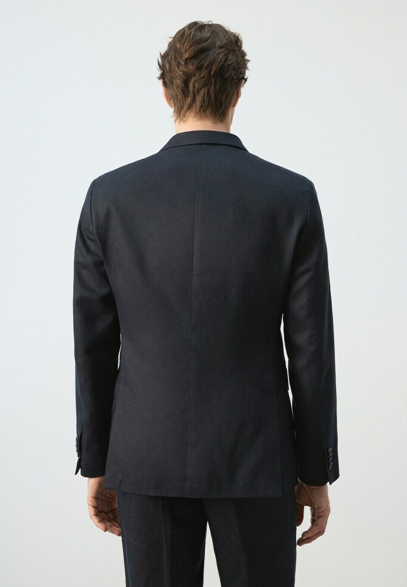 Homme debout, de dos, portant une veste de costume noire ajustée et un pantalon assorti, sur un fond clair uni.