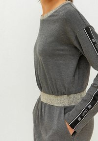 Jumpsuit gris à manches longues avec une taille cintrée, doté d'une ceinture élastique métallique et de bandes latérales noires contrastantes avec des accents réfléchissants.