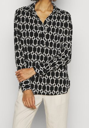 Blouse noire à manches longues avec motif de chaîne blanche entrelacée, portée avec un pantalon de couleur claire, mains détendues au niveau de la taille.