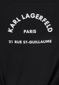 Juodas audinys su baltais užrašais "KARL LAGERFELD," "PARIS," ir "21 RUE ST-GUILLAUME" išlenktame išdėstyme. Lygus ir juostuotas tekstūros.