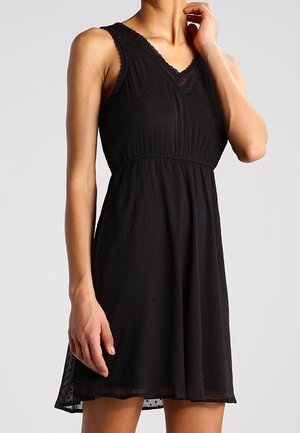 Robe noire sans manches avec bordure en dentelle sur l'encolure en V, taille cintrée et jupe légèrement évasée portée par une personne sur un fond uni.