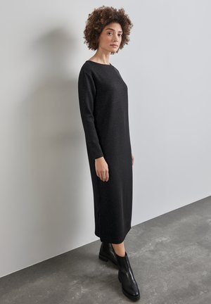 Schwarzes langärmliges Kleid mit geradem Schnitt und leichten Seitenschlitzen. Hergestellt aus strukturiertem Stoff mit dezenten Glitzerpartikeln. Kombiniert mit schwarzen Knöchelstiefeln.