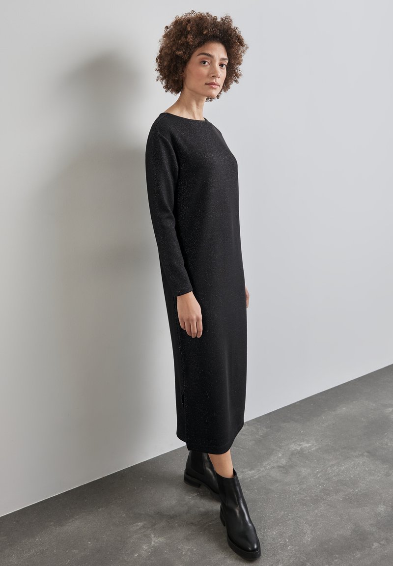 Robe noire à manches longues avec une coupe droite et de légères fentes sur les côtés. Fabriquée dans un tissu texturé avec des éclats discrets. Portée avec des bottines noires.