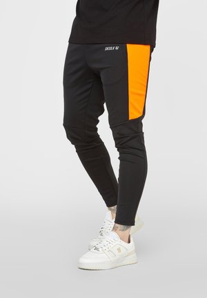 Zwarte broek met een slanke pasvorm, met feloranje zijpanelen en witte sneakers, gedragen door een persoon die staat tegen een effen achtergrond.