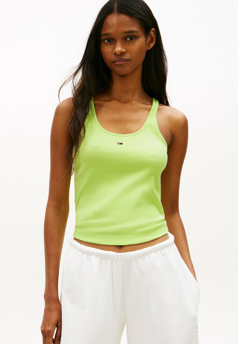 Frau mit langen dunklen Haaren, die ein ärmelloses, limettengrünes Crop-Top und weiße Hosen mit elastischem Bund trägt, vor einem einfarbigen Hintergrund stehend.