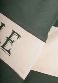 Zielono-kremowa, w paski, bawełniana tkanina z wyraźnym napisem na zielonej części. Tekstura wydaje się miękka i gładka.