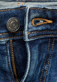 Jeans de mezclilla azul oscuro con una cremallera visible, botón de metal con grabado "MNG DENIM" y costuras de contraste en marrón dorado.