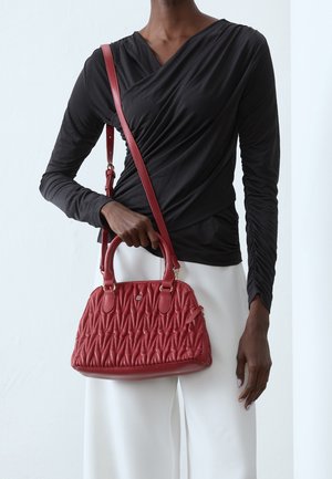 Person iført en sort ruched langærmet top og hvide bukser, der holder en tekstureret rød læderhåndtaske med både håndtag og skulderrem.