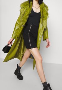 Femme marchant portant un manteau à col en fausse fourrure verte, une robe noire avec fermeture éclair devant, des bottines noires et portant un sac à main noir.