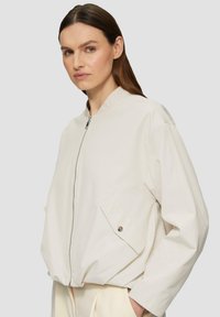 Veste bomber zippée beige clair avec un col rond, des poches latérales et une coupe décontractée. Tissu doux et lisse avec un détail de couture minimal.
