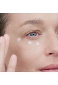 Vit kräm appliceras under ögat, åtföljt av en hand med polerade, naturliga naglar. Huden är slät med en naturlig ton och struktur.