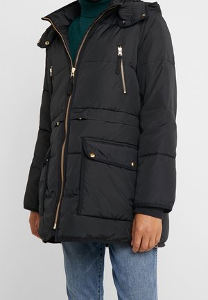 Zwarte gewatteerde jas met gouden ritsen en knopen, gedragen over een groene coltrui en gecombineerd met blauwe spijkerbroek.