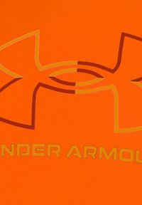 Una tela deportiva de color naranja brillante presenta un gran logo estilizado con un contorno dorado, acompañado de un texto dorado que dice "UNDER ARMOUR".