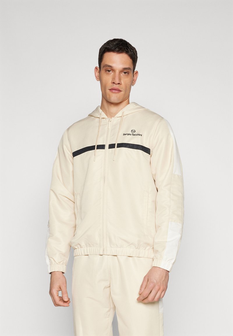 Chaqueta con capucha beige con cremallera y una franja negra en el pecho, puños elásticos y logo en el lado izquierdo. Textura de tela suave.