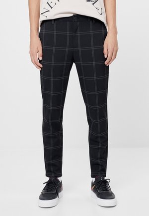 Pantalones ajustados negros con patrón de cuadros tipo ventana en gris claro, combinados con zapatillas negras con suelas blancas y una camiseta gráfica blanca metida por dentro.