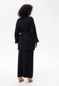 Czarny kardigan z paskiem, z długimi, szerokimi rękawami, nałożony na długą czarną spódnicę, połączony z beżowymi butami na wysokim obcasie. Teksturowana tkanina.