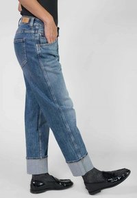Persona vistiendo jeans bicolor azul con puños, calcetines oscuros y mocasines negros brillantes, de pie con una mano en el bolsillo frente a un fondo liso.