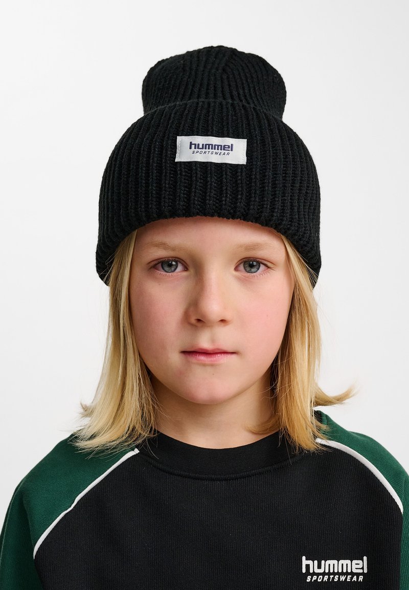 Svart ribbad mössa med uppvikt kant och en vit etikett med "hummel Sportswear". Bärs med en svart och grön sweatshirt.