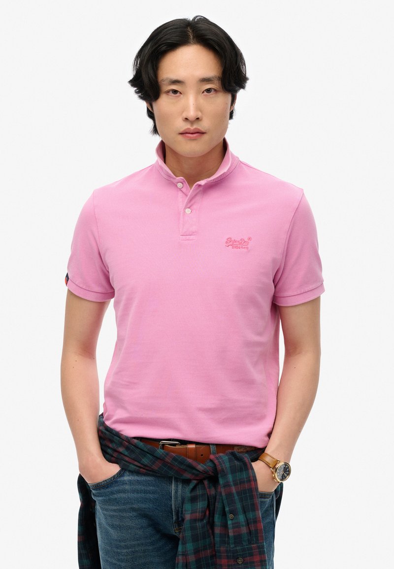Lyserød polo-shirt med krave og to knapper, med broderet logo. Bæres med blå jeans, brun bælte og en ternet skjorte bundet omkring taljen.