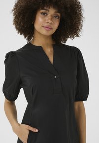 Crna haljina s kratkim puff rukavima, V-izrezom i prednjim dugmadi. Glatka tkanina s opuštenim krojem i bočnim džepovima.