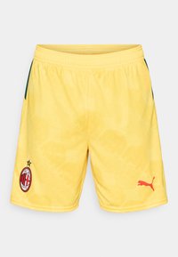 Gule idrettsshorts med et teksturert mønster, som har en rød Puma-logo og AC Milan-emblemet med en stjernedetalj på venstre ben.