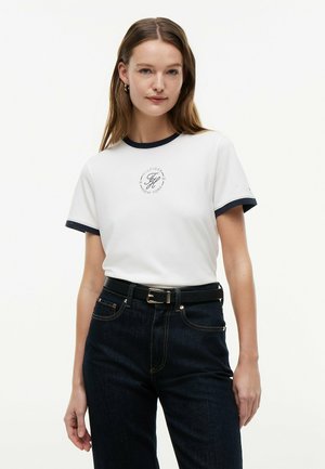 Kobieta w białym T-shircie z granatowymi lamówkami i logo, w połączeniu z ciemnymi jeansami z wysokim stanem i czarnym paskiem, stojąca na jednolitym tle.