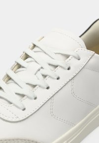 Zapatilla de cuero blanco con cordones planos blancos, detalles cosidos, una superficie suave y un diseño de perfil bajo con una suela de goma.