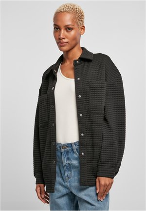 Black quilted shirt jacket with snap buttons, two chest pockets, and a relaxed fit, worn over a white tank top and blue high-waisted jeans.