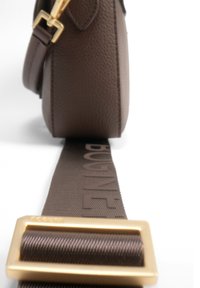 Brun lædertaske med en struktureret overflade, der har en bred brun rem med prægede bogstaver og en spænde i guldfarvet metal.