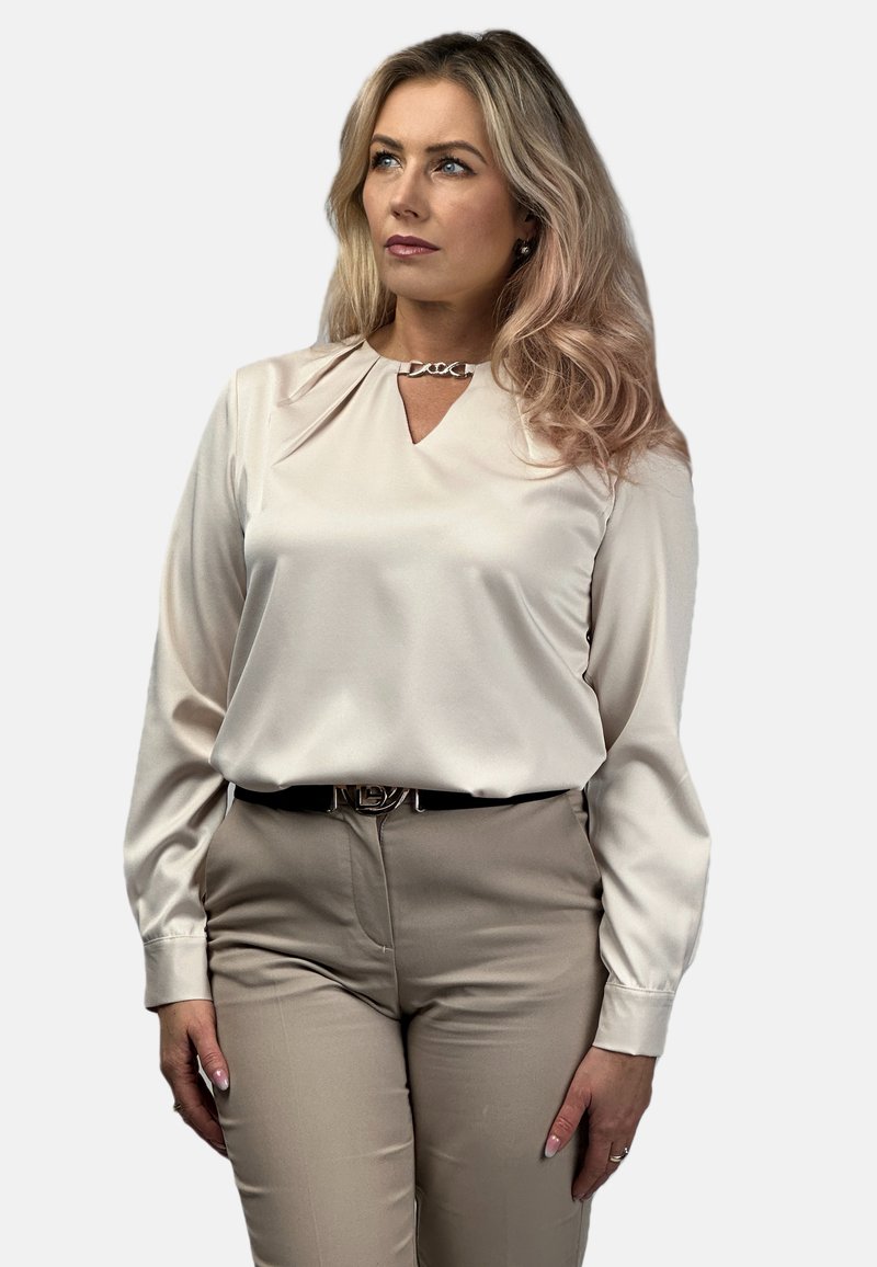 Kremowa bluzka z dekoltem w serek i srebrnym łańcuszkiem przy kołnierzy, zestawiona z beżowymi spodniami. Gładki materiał z długimi rękawami.