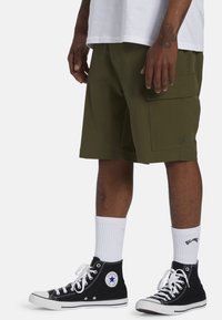 Les shorts cargo en vert olive présentent une grande poche latérale, associés à des baskets montantes noires et des chaussettes blanches. Tissu texturé visible.