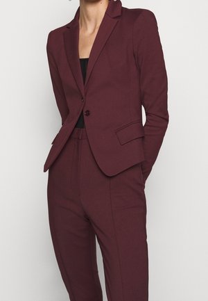 Femme portant un blazer bordeaux ajusté avec un pantalon assorti et un haut noir, debout les mains dans les poches devant un fond uni.