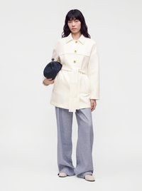 Femme portant une veste blanche ceinturée, un pantalon large clair, tenant une pochette noire texturée sur un fond uni.