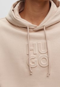 Sudadera beige con un logotipo texturizado "HUGO" en la parte frontal, con capucha ajustable y un tejido suave.